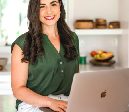 brown haired girl sitting in the kitchen on her computer wearing a green shirt and white pants